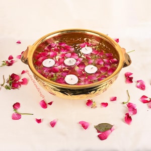 Könnte beinhalten: Eine goldene Metallschale gefüllt mit Wasser und Rosenblättern, mit schwimmenden Teelichtern. Die Schale hat dekorative Gravuren und zwei Griffe.
