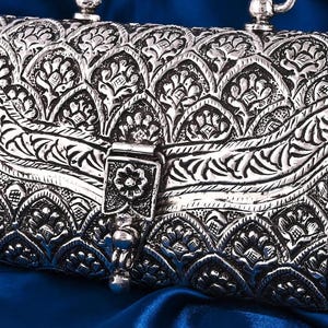 May include: A silver-colored, rectangular handbag with intricate floral and leaf patterns. The bag features a clasp closure and a chain strap. The background is a deep blue fabric, creating a contrast with the metallic finish of the purse.