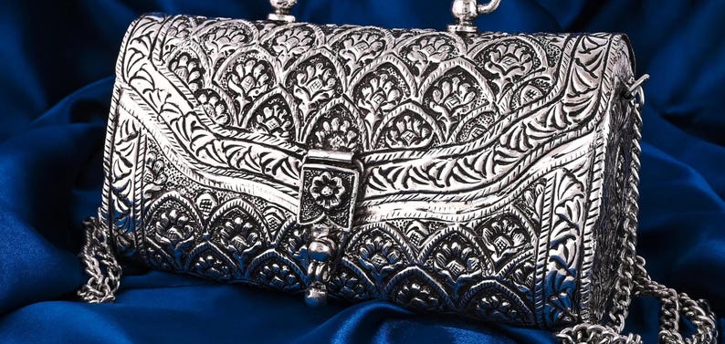May include: A silver-colored, rectangular handbag with intricate floral and leaf patterns. The bag features a clasp closure and a chain strap. The background is a deep blue fabric, creating a contrast with the metallic finish of the purse.