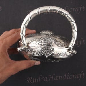 May include: A silver, round, decorative box with an ornate design and a handle. The box is closed and has a detailed, intricate pattern.