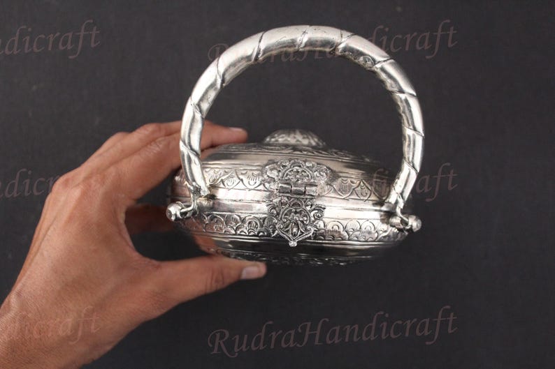 May include: A silver, round, decorative box with an ornate design and a handle. The box is closed and has a detailed, intricate pattern.