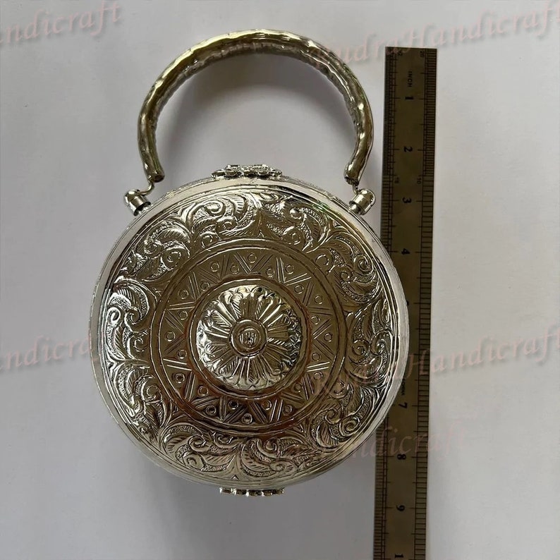 May include: A silver, round, decorative box with an ornate design and a hinged lid. The box has a silver handle and is approximately 4 inches in diameter.