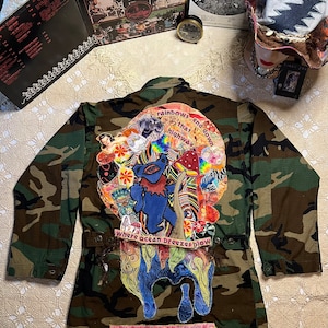 May include: A camouflage jacket with a large, colorful patch on the back. The patch features a cartoon bear, flowers, mushrooms, and the text "rainbows and down that highway" and "Where ocean breezes blow".