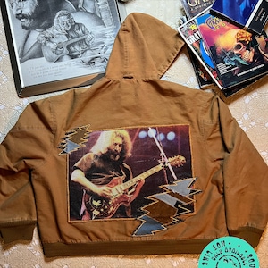 May include: A brown hooded jacket with a large embroidered patch on the back featuring a musician playing a guitar. The patch is surrounded by a jagged lightning bolt design. The jacket has a brown ribbed knit collar, cuffs, and waistband. The jacket has a small circular patch on the bottom right corner with the text "Other Girls - Soul Threads - Only Available Thru"