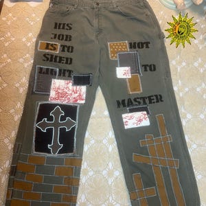 May include: Olive green work pants with various fabric patches and text. The text reads "HIS JOB IS TO SHED LIGHT NOT TO MASTER." Patches include a cross, brick pattern, and patterned fabrics. The pants have a relaxed fit.
