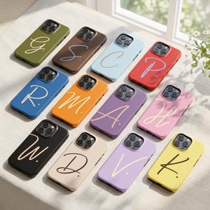 May include: A selection of mobile phone cases in a variety of colours, each displaying a different initial in a cursive font. The cases are in shades such as green, brown, blue, orange, purple, pink, black, beige, lavender, and yellow. They are arranged on a white surface.