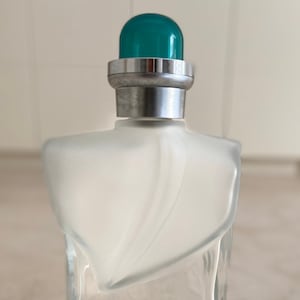 May include: A clear glass bottle of perfume with a green and silver cap. The bottle is labeled "Noa Noa" and "Otto Kern".
