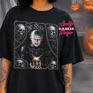 May include: A black short-sleeve t-shirt featuring a graphic print of a pale, pin-headed figure holding a glowing puzzle box, surrounded by four skulls and chain motifs. The text Loulife TUMBLER Designs is printed in pink on the upper right side.