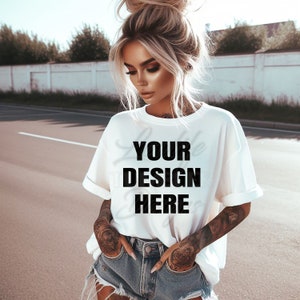May include: A white oversized t-shirt with the text "YOUR DESIGN HERE" printed on the front. The shirt is being worn by a person with blonde hair and tattoos on their arms.