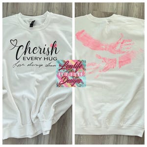 May include: White sweatshirt with the words "Cherish Every Hug" in black script and a heart graphic. The back of the sweatshirt features a pink handprint design. The sweatshirt is from Loulife Tumbler Designs.