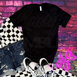 May include: A black t-shirt, denim shorts, and black and white checkered sneakers are arranged on a black and white checkered blanket. The background is a brick wall with a neon pink balloon dog.
