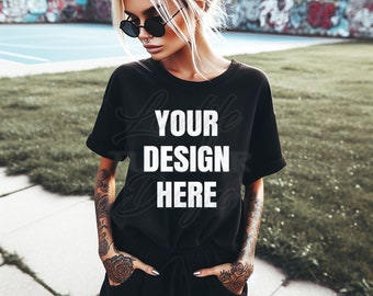 Op de afbeelding: Een zwart T-shirt met de tekst "YOUR DESIGN HERE" in wit gedrukt op de voorkant. Het T-shirt wordt gedragen door een persoon met tatoeages op hun armen.