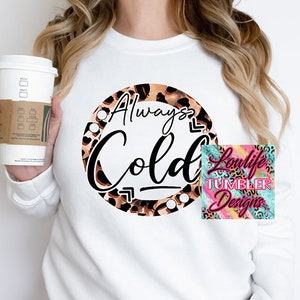 Op de afbeelding: Witte sweatshirt met een cirkelvormig luipaardprintontwerp met de tekst "Always Cold" in zwart schrift. Een koffiekopje wordt in de afbeelding vastgehouden. Een klein logo voor "Loulife Tumbler Designs" staat rechtsonder.