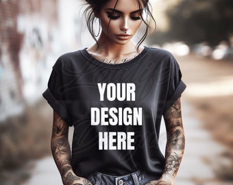 Op de afbeelding: Een zwart T-shirt met de tekst "YOUR DESIGN HERE" in wit gedrukt op de voorkant. Het shirt wordt gedragen door een persoon met tatoeages op hun armen.