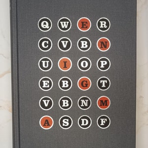 May include: A dark gray book with a grid of letters in white circles, some with orange centers. The letters are arranged in a grid pattern. The book's cover has a textured appearance.