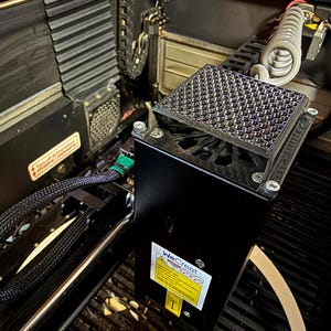 May include: Close-up of a black laser cutter machine with a textured metal surface and a black box with a label that says "WeCreate". The machine has a green cable and a fan.