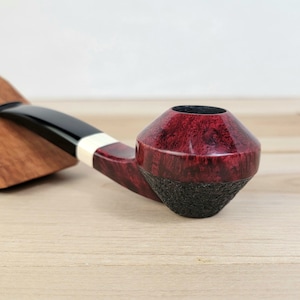 May include: A red and black briar pipe with a rounded bowl and a black stem. The pipe is resting on a wooden surface.