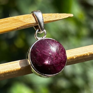 Pode incluir: Um pendente redondo, vermelho escuro, com uma argola prateada. O pendente é segurado por dois palitos de madeira contra um fundo verde desfocado. O pendente tem uma superfície polida e reflexiva.