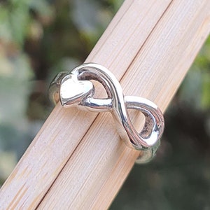 May include: A silver infinity ring with a small heart charm. The ring is on a wooden surface.