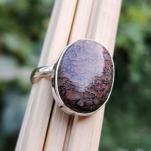 Pode incluir: Um anel de prata com uma grande pedra oval de osso de dinossauro fossilizado marrom e vermelho.