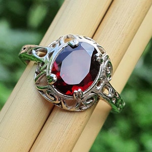 Puede incluir: Un anillo de plata con un diseño adornado presenta una gran piedra roja de forma ovalada.