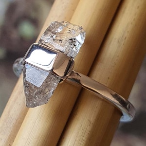 May include: A silver ring featuring a clear, faceted, raw diamond crystal set in the centre. The diamond is a pointed, irregular shape.