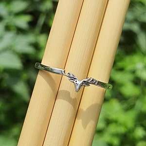 Könnte beinhalten: Ein silberfarbener Ring mit einem Hirschgeweih-Design. Der Ring ist dünn und filigran, mit dem Geweih-Design in der Mitte des Bandes. Der Hintergrund ist ein verschwommenes Grün, was auf eine Umgebung im Freien hindeutet. Der Ring wird auf einer hellbraunen Oberfläche präsentiert.