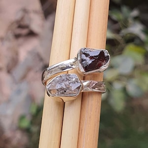 May include: A silver ring with two raw gemstones. The larger stone is a clear, faceted crystal. The smaller stone is a dark red, rough crystal.