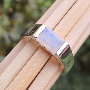 Puede incluir: Un anillo de plata con una piedra de luna rectangular engastada en el centro. El anillo está sobre una superficie de madera.