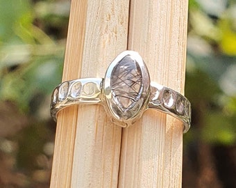 Op de afbeelding: Een zilveren ring met een ovale, heldere edelsteen met zwarte insluitsels. De band heeft een gehamerde textuur met cirkelvormige inkepingen. De ring wordt gepresenteerd op een houten oppervlak, met een vage groene achtergrond.