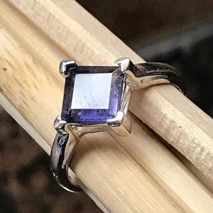 Pode incluir: Um anel de prata com uma pedra preciosa roxa intensa de corte quadrado fixada por quatro garras. A banda &eacute; de prata polida e a pedra apresenta uma superf&iacute;cie plana e reflexiva com reflexos de luz vis&iacute;veis na sua faceta superior.
