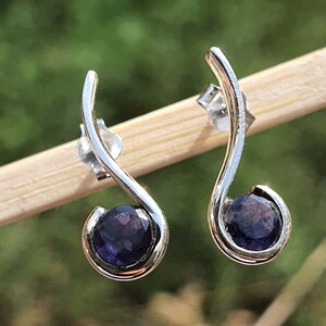 Peut inclure: Une paire de boucles d'oreilles en argent avec un design incurvé et une pierre précieuse ronde de couleur violet foncé dans chaque boucle d'oreille.