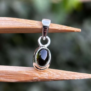 May include: A silver pendant featuring a smooth, oval-shaped dark green gemstone set in a polished metal bezel. The pendant includes a simple silver bail at the top for attachment to a chain, showcasing a clean and reflective metallic finish.