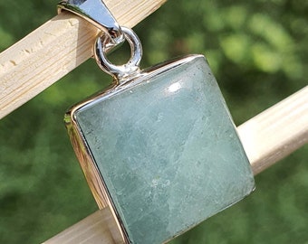 May include: A silver pendant with a square aquamarine gemstone. The pendant is on a wooden surface.