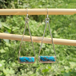 Puede incluir: Un par de pendientes triangulares de plata con detalles de piedra turquesa. Los pendientes están colgados de una clavija de madera.