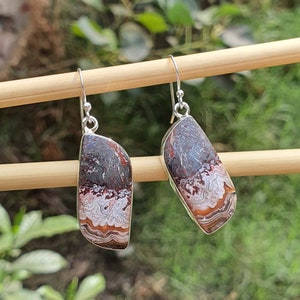 May include: A pair of silver-tone earrings with a unique stone design. The earrings feature a rectangular shape with a natural stone pattern in shades of gray, red, and brown. The stone is set in a silver frame with a hook closure.