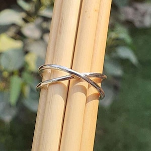 May include: A polished silver-toned ring featuring a crossover design with two intersecting bands that form an X shape. The ring is positioned around two smooth, light-colored wooden dowels, showcasing its metallic finish and curved structure.