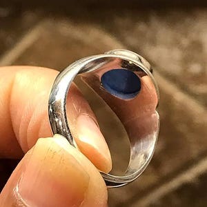Puede incluir: Un anillo de plata con una banda de metal lisa y pulida, y una piedra circular de color azul intenso engastada en la cabeza de la pieza. El interior de la banda del anillo es visible, mostrando una superficie met&aacute;lica reflectante y la parte inferior de la piedra.
