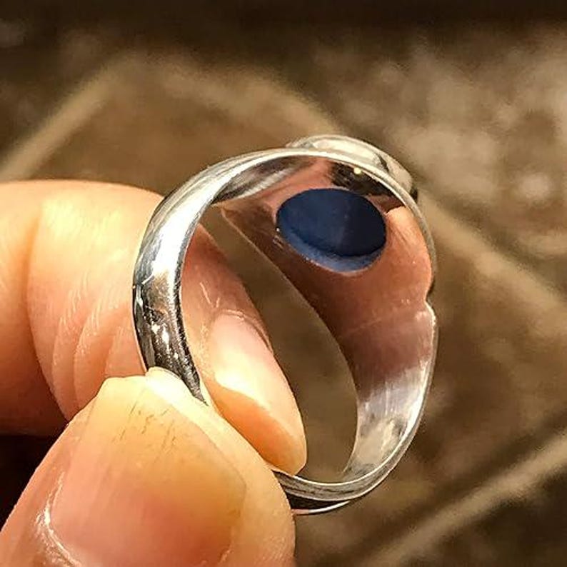 Puede incluir: Un anillo de plata con una banda de metal lisa y pulida, y una piedra circular de color azul intenso engastada en la cabeza de la pieza. El interior de la banda del anillo es visible, mostrando una superficie met&aacute;lica reflectante y la parte inferior de la piedra.