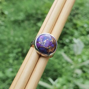May include: A silver ring with a round purple and gold gemstone. The ring is set on a wooden surface.
