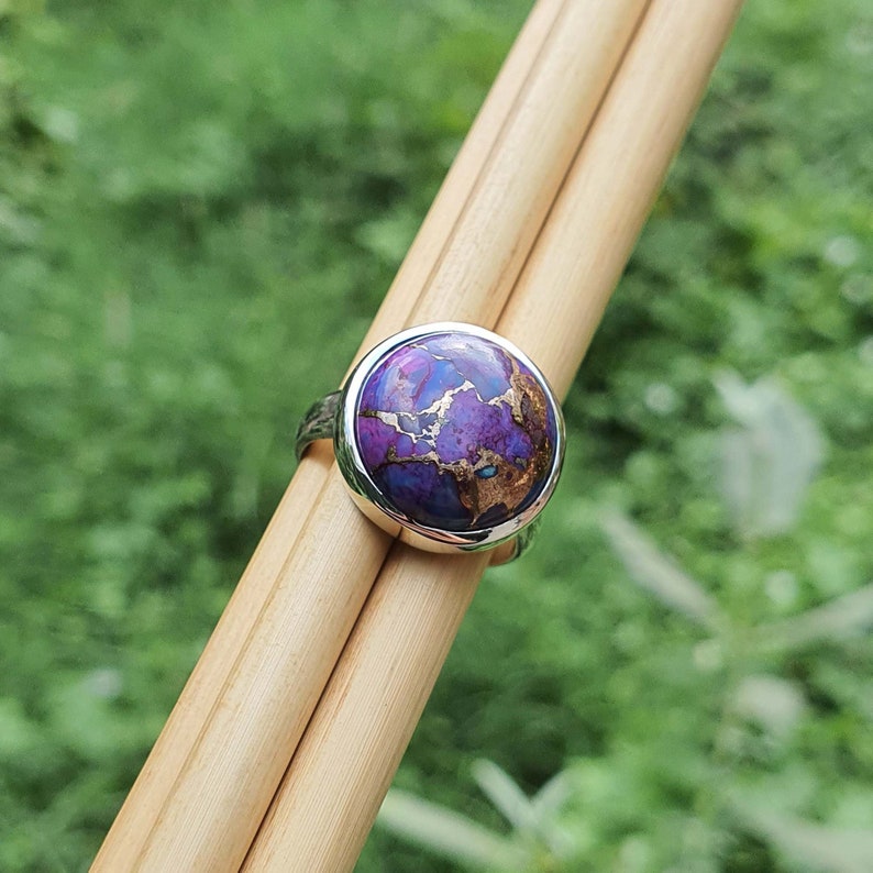 May include: A silver ring with a round purple and gold gemstone. The ring is set on a wooden surface.