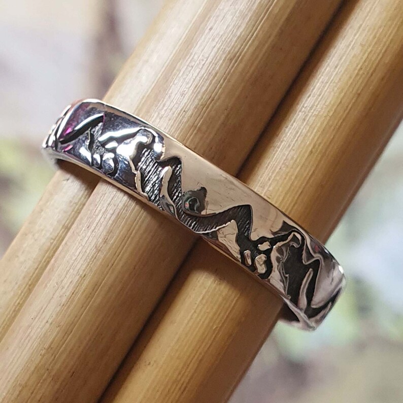 May include: A silver ring with a textured design of a mountain range. The ring is on a wooden surface.