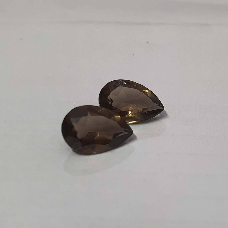 May include: Two pear-shaped smoky quartz gemstones. The gemstones are a dark brown color and have a faceted cut.