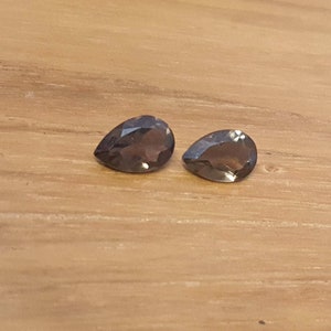 May include: Two pear-shaped, dark gray gemstones on a wooden surface.
