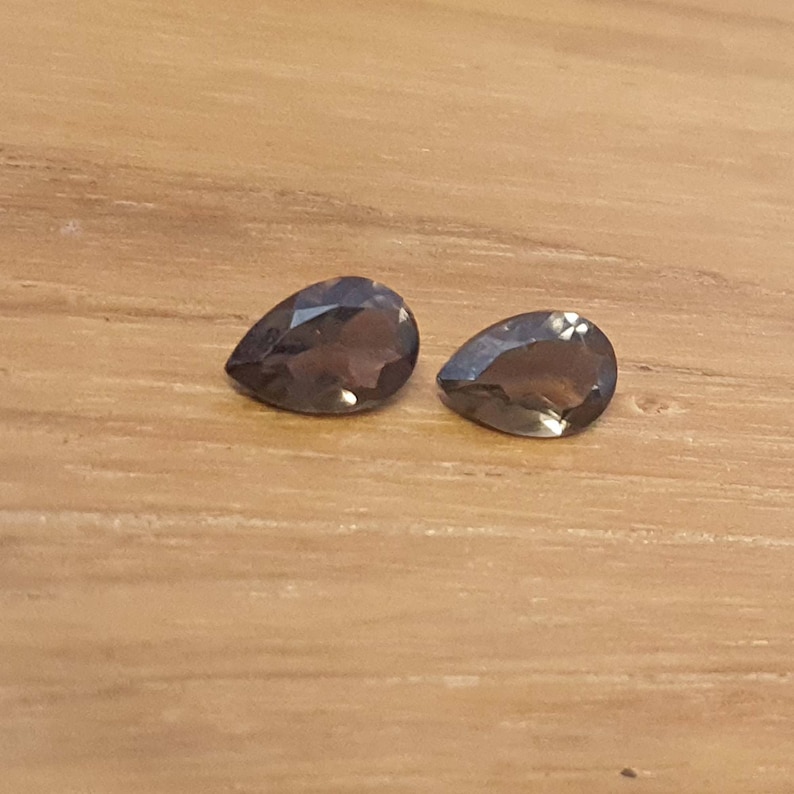 May include: Two pear-shaped, dark gray gemstones on a wooden surface.