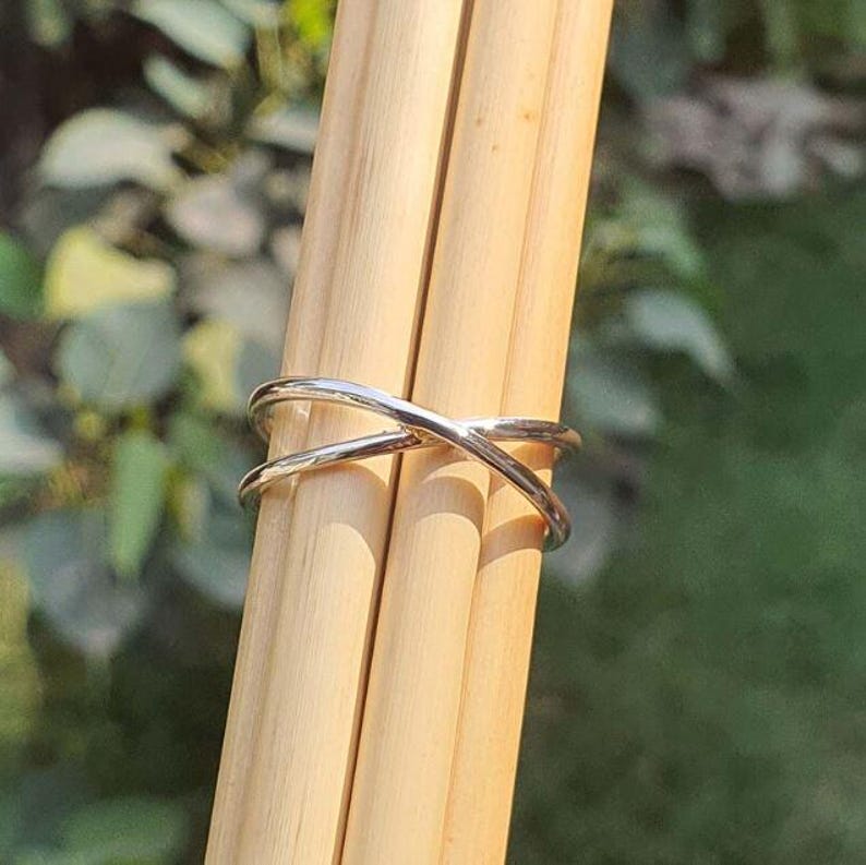 May include: A polished silver-toned ring featuring a crossover design where two thin bands intersect to form an X shape. The ring is positioned horizontally around three light-colored wooden dowels, showcasing its smooth, reflective metallic surface.