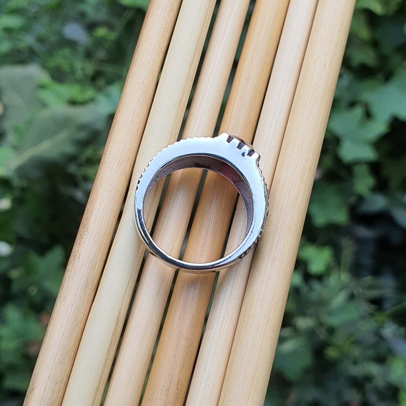 May include: A silver ring with a row of small clear stones around the band. The ring is on a background of wooden sticks.