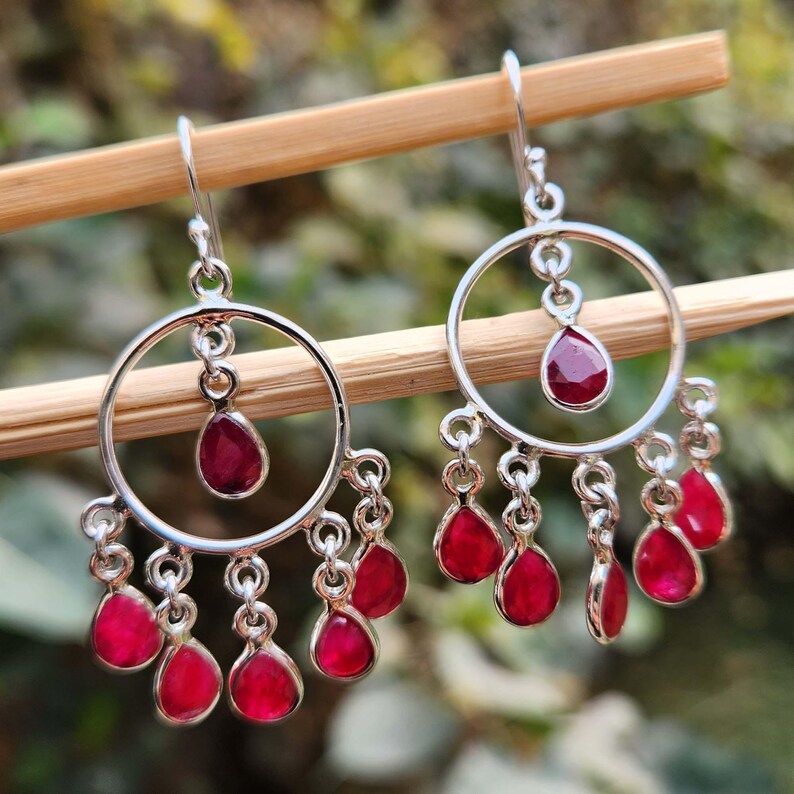 May include: Silver hoop earrings with dangling red ruby stones. The earrings have a circular design with a single ruby stone at the top and multiple ruby stones dangling from the bottom.