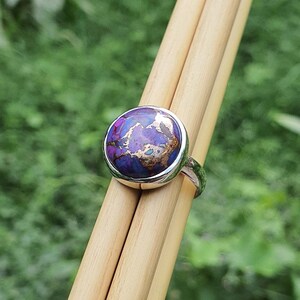 May include: A silver ring with a round purple and gold gemstone. The ring is set on a wooden surface.