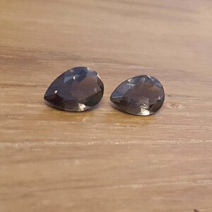 May include: Two pear-shaped, dark gray gemstones on a wooden surface.